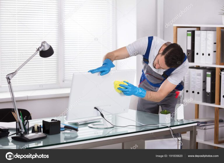 Young Male Janitor Cleaning Computer With Sponge In Office